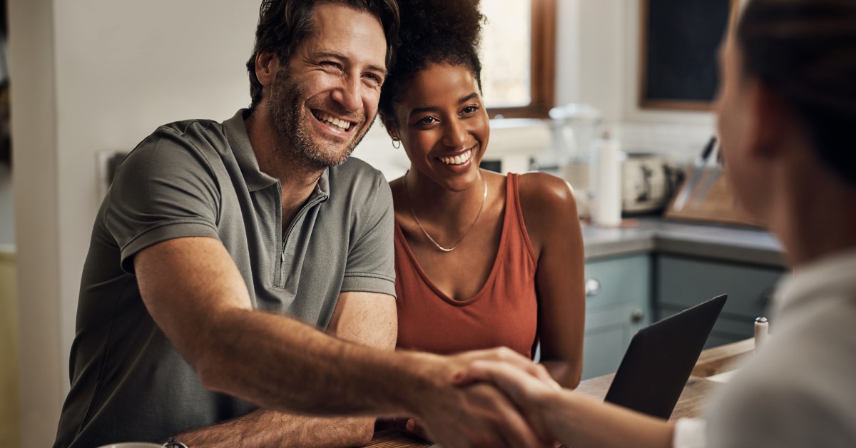 Two people shake hands over a wood table. One person is smiling, and there is another person smiling next to them.