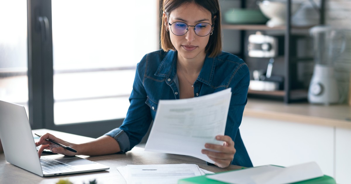 A person wearing round-rimmed glasses and a blue jacket, reading documents in front of a computer in their kitchen.