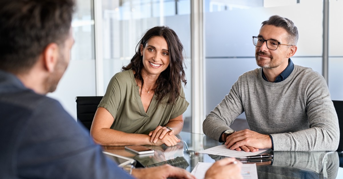 Two people sitting in a large, windowed office, smiling at their financial advisor. They each have papers in front of them.