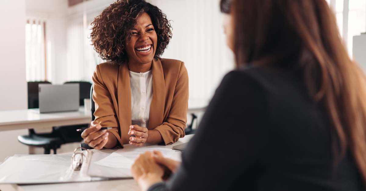 A woman wearing a brown suit is laughing while sitting in an office and looking at a woman sitting across from her.