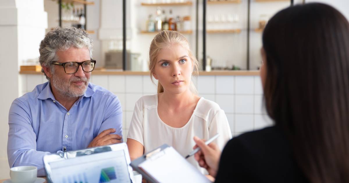 A couple sitting down in their kitchen as a financial expert talks to them while using a clipboard and laptop.