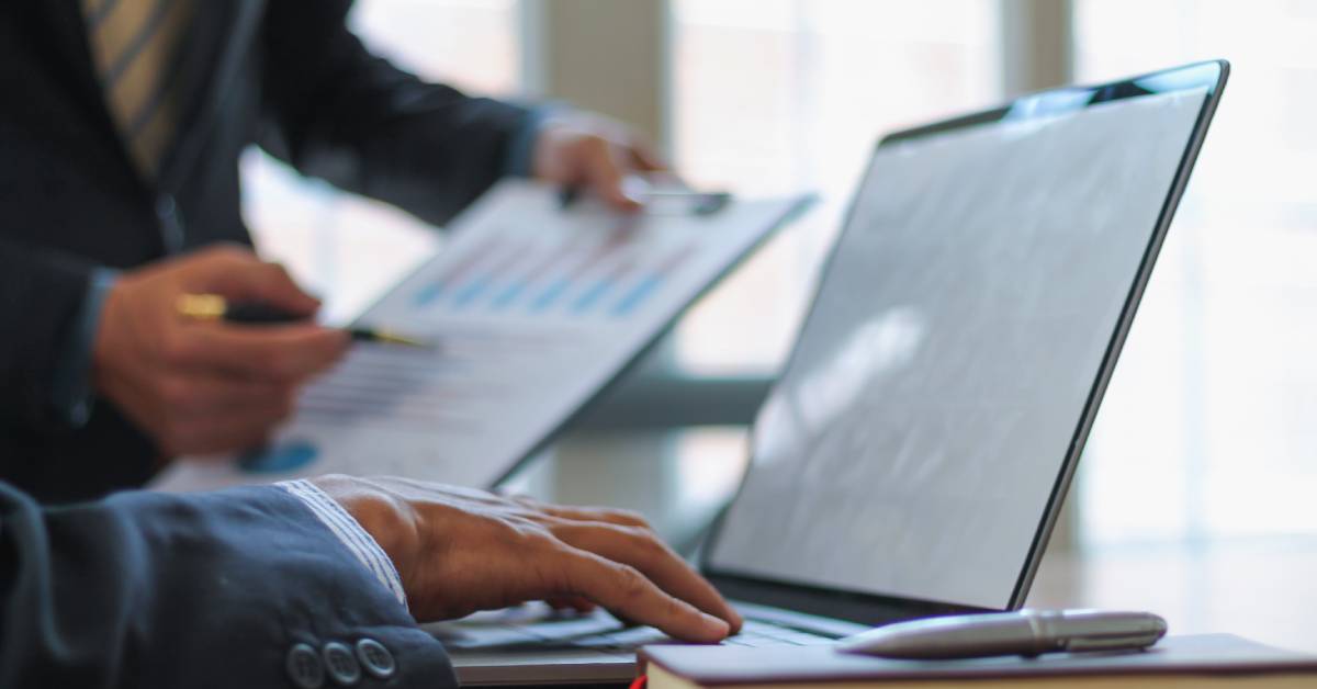 A person gesturing to a clipboard and standing next to another person who uses a laptop that sits next to a book and pen.