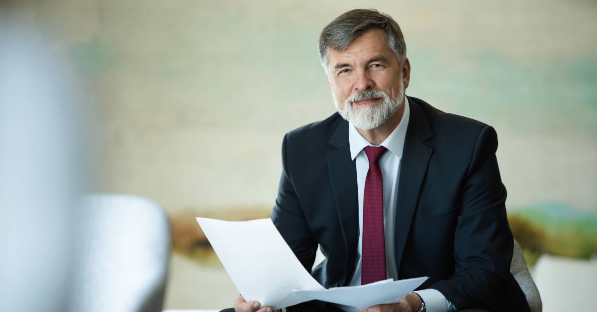 A person in a dark-colored suit and red tie sitting on a couch and holding multiple documents. He smiles at the camera.