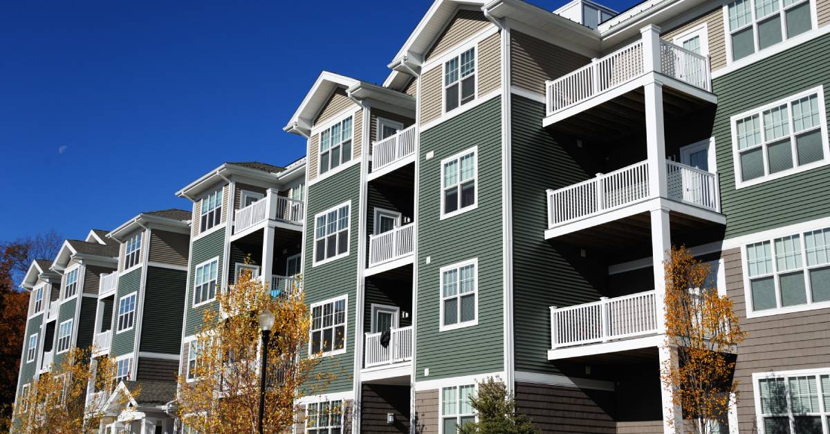 A multi-colored apartment building has white railings on each floor and several trees near the entrance.