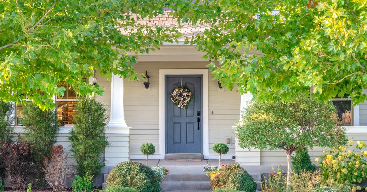 A cream-colored house has a dark blue door. The lawn has vibrant green grass, trees, and bushes on it.