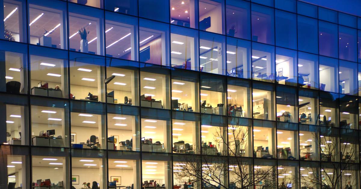 A massive office building is full of workers and bright lights that make it glow in the dark evening.