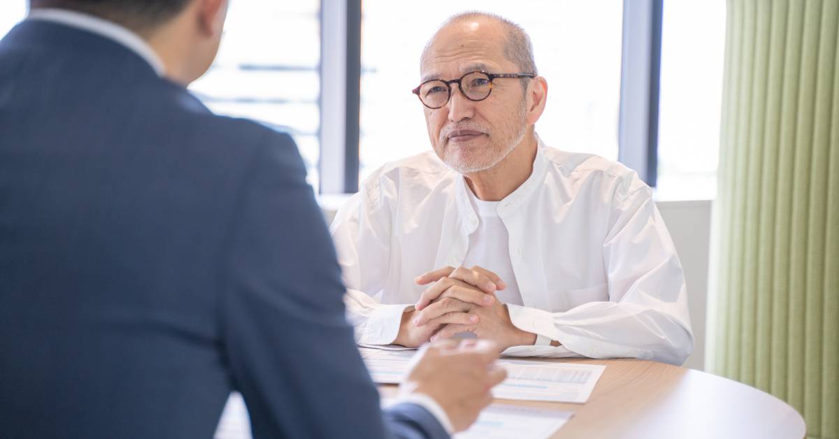 An elderly person wearing round-rimmed glasses is sitting at a table and listening to someone sitting across from him.