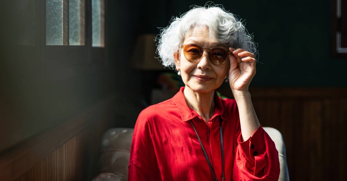 A person with white hair and a red shirt is putting their left hand on their dark round-rimmed glasses.