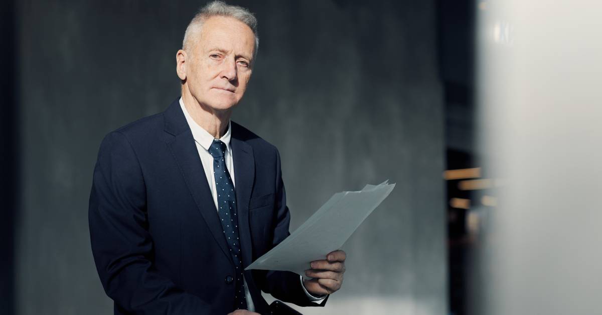 An elderly person wearing a high-end suit is holding paper documents and a cell phone in their hands.