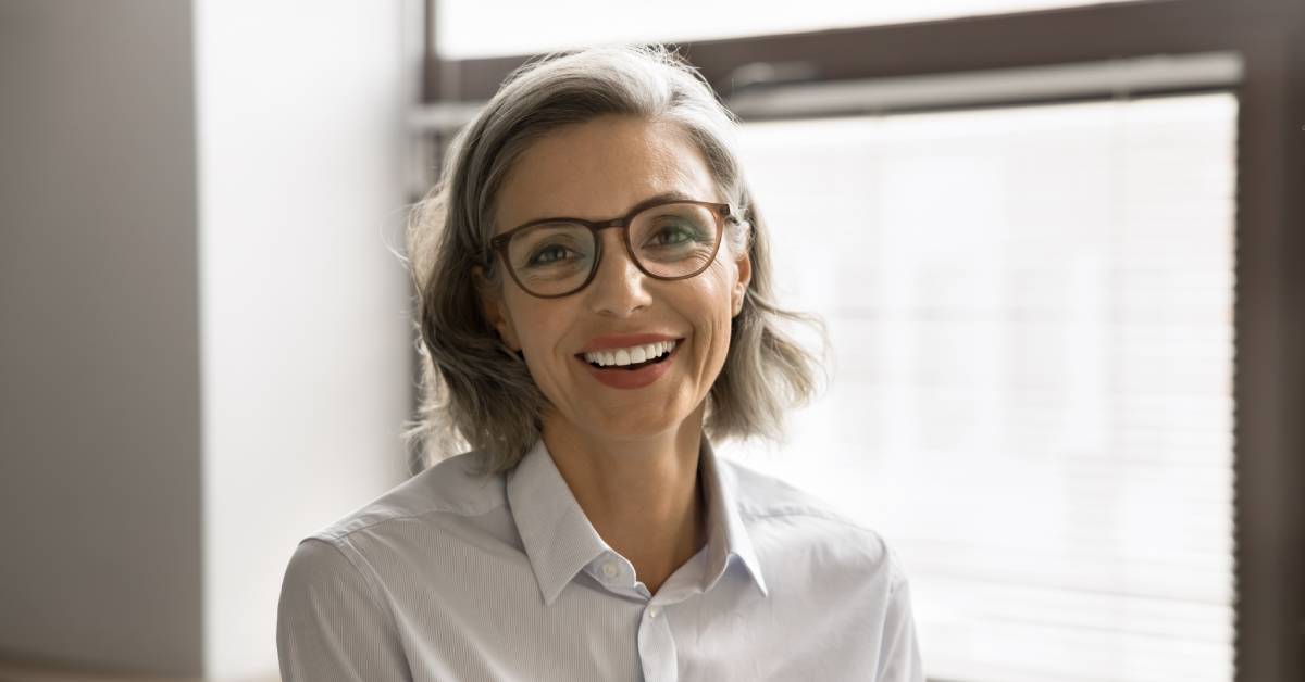 A person wearing brown glasses and a white shirt is sitting alone in front of a window and smiling.