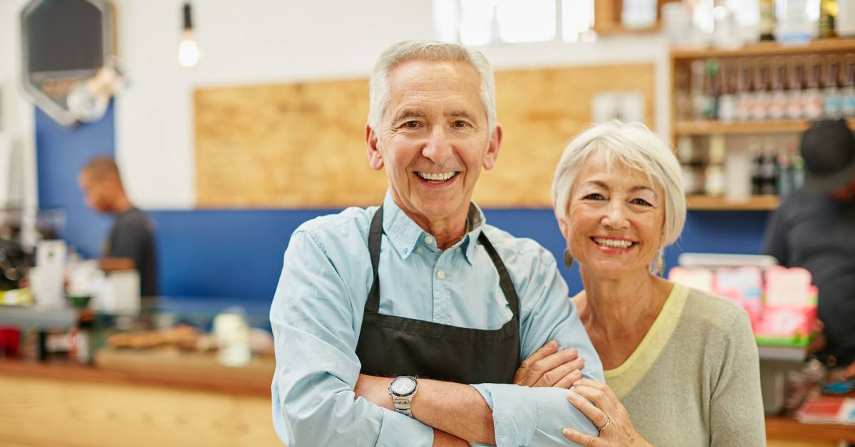 Two people are standing and smiling in the middle of a shop. Behind them is a counter where two employees are working.