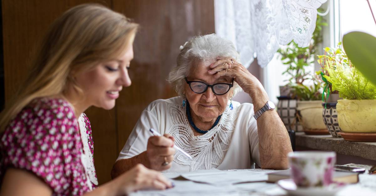 Two people sitting at a table. The table is covered in paperwork, as well as a small book and a teacup.