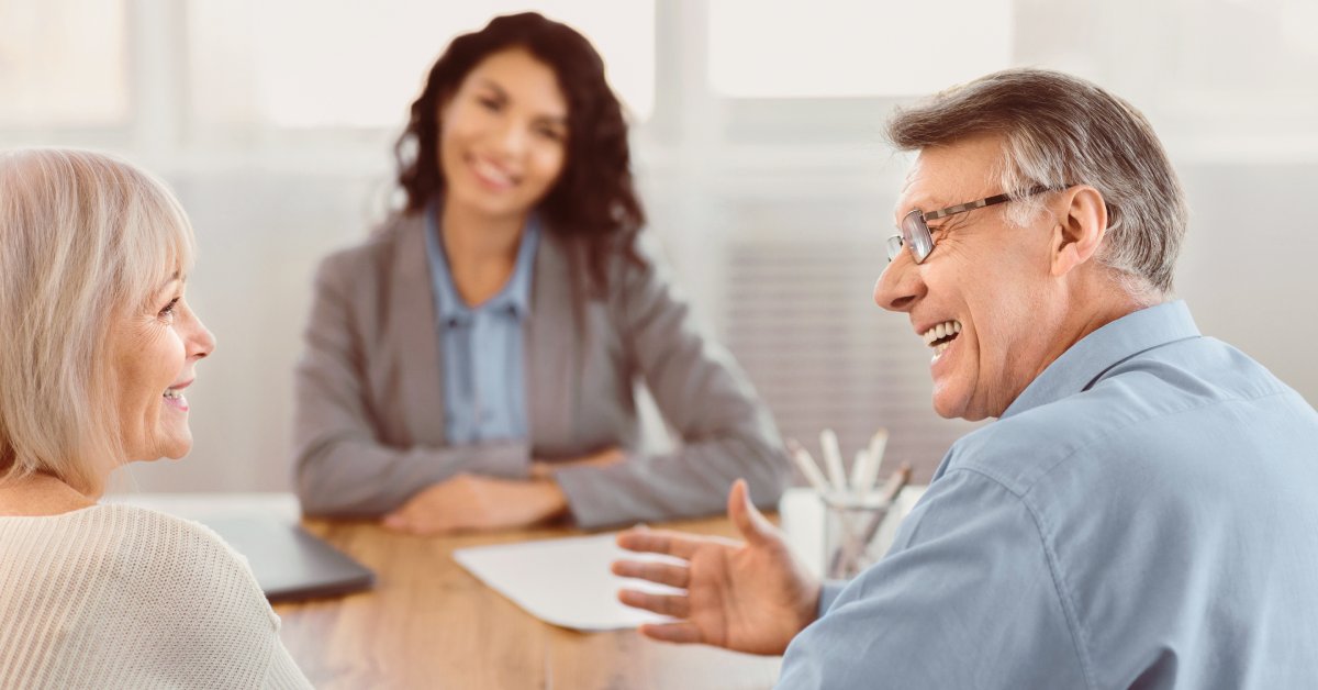 Two people sitting at a table and smiling at each other. Another person sits across from them wearing business attire.