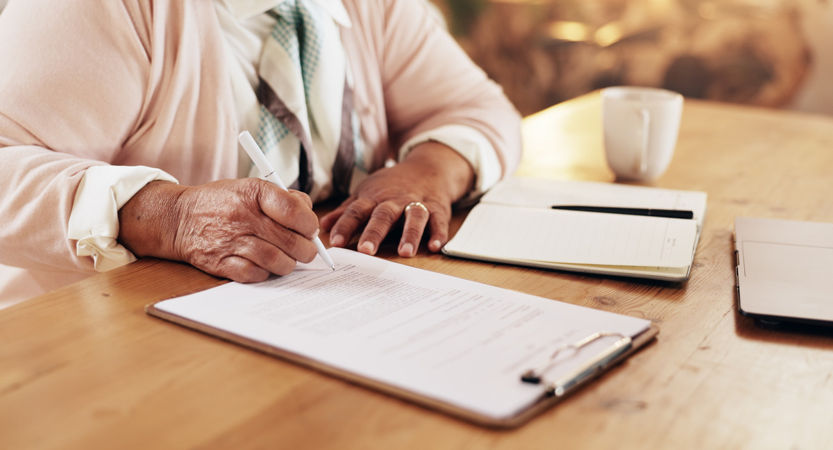 A person using a white pen to write on a document attached to a clipboard. The clipboard is on a wooden table.