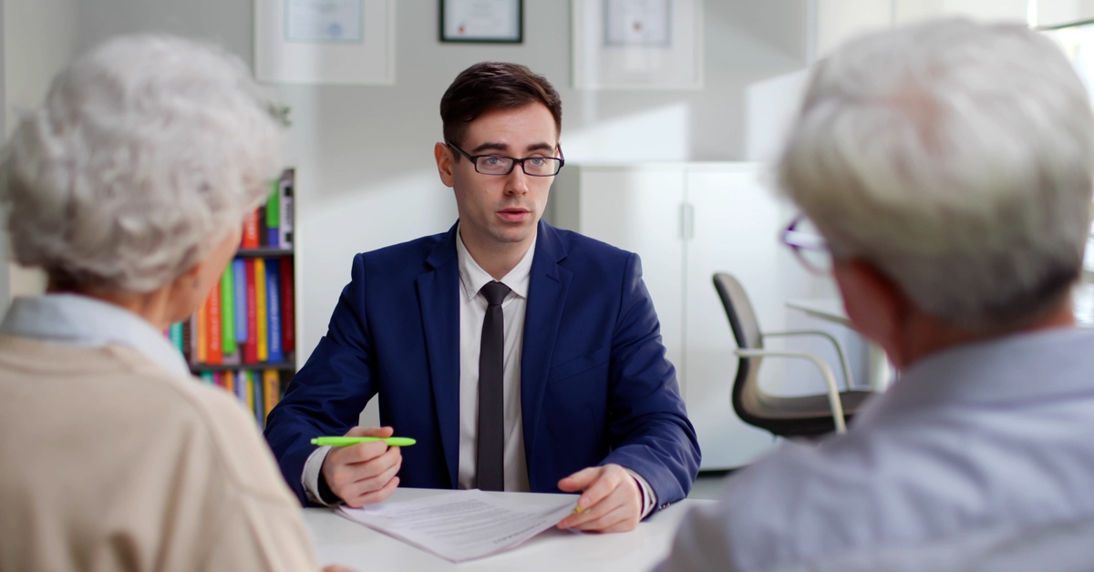 A person wearing glasses and a blue business suit sits at a table while talking to two people sitting across the table.