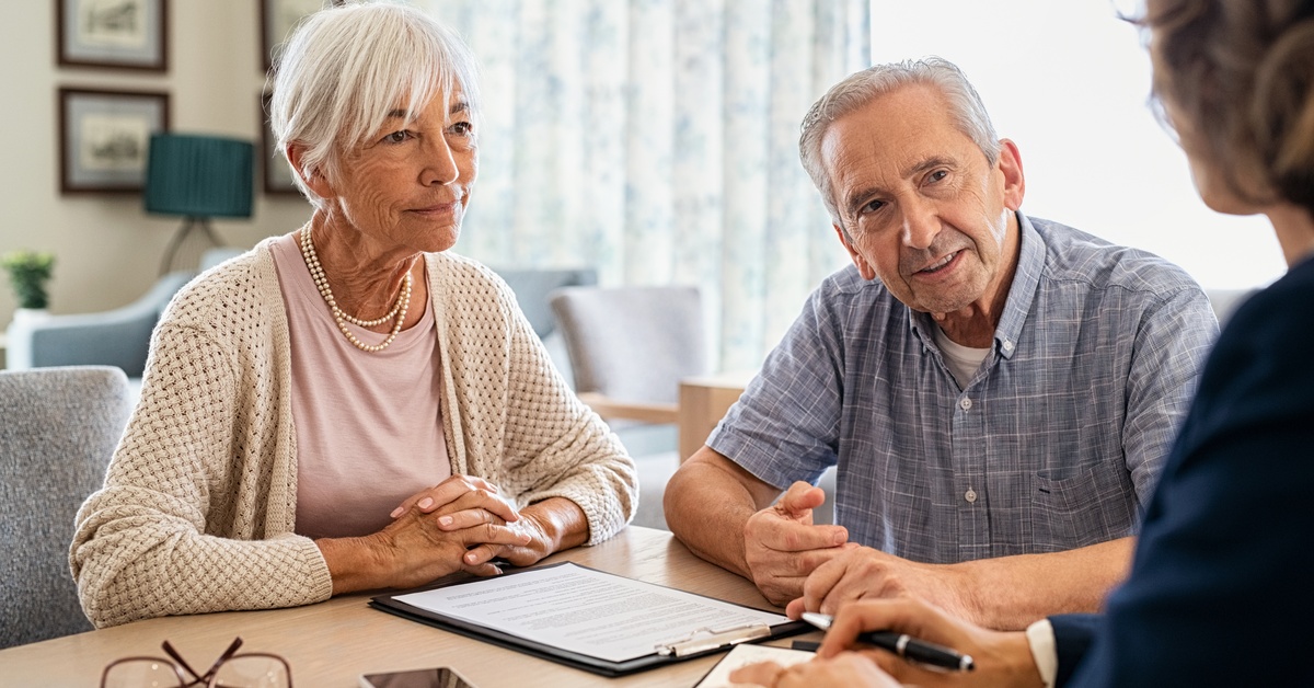 A couple sitting at a wooden table, looking at another person who is talking to them and holding a pen.
