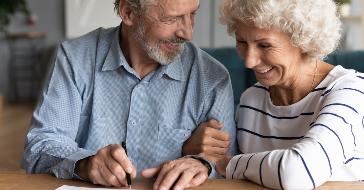 An older couple sitting at a table and smiling at each other as one prepares to sign a paper document with a pen.