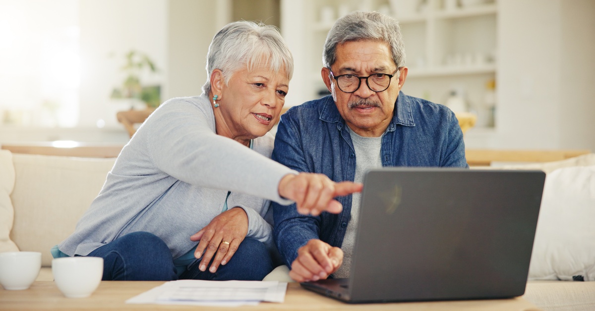 Two people sitting on a couch. One of the people is pointing at a laptop that is on the table in front of them.