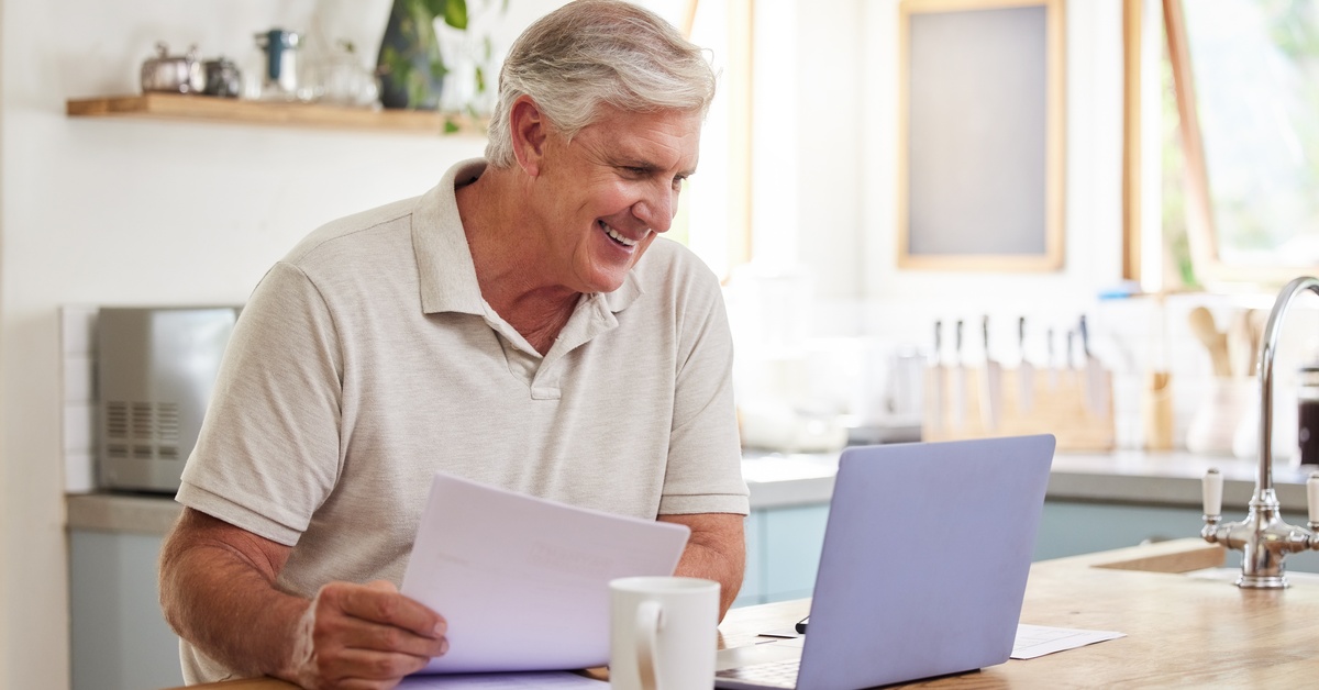 A person is smiling while sitting down at a wooden countertop. They are holding documents while looking at a laptop.