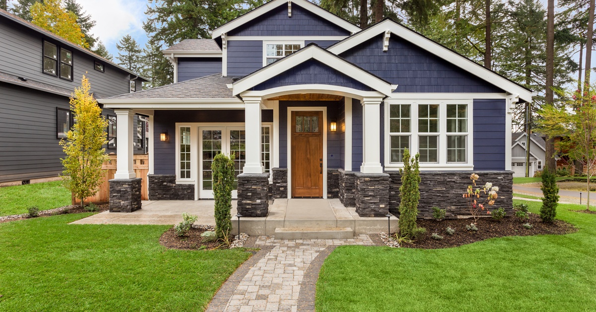 A large blue home in a neighborhood during the day. The house has two lights outside that are illuminated.
