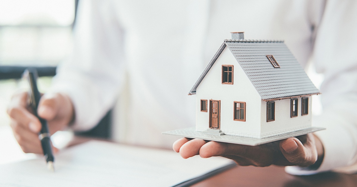 A person sitting at a desk and signing a document with their right hand. A small model of a house is in their left hand.