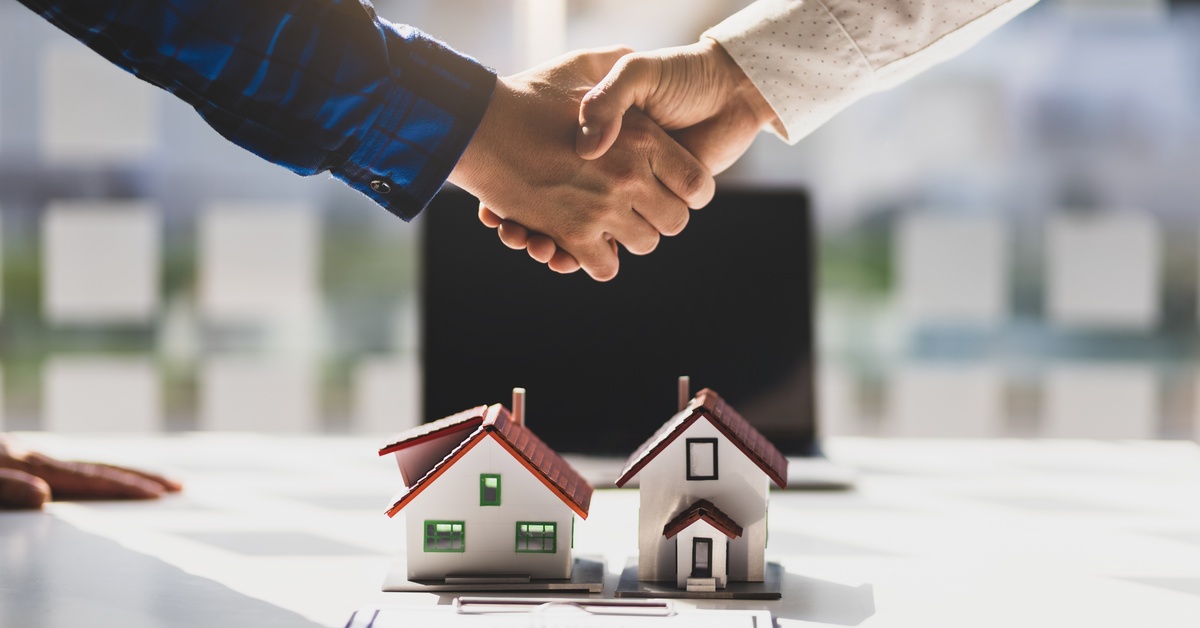 Two people are shaking hands over a table. The table has a clipboard and two small models of homes on top of it.