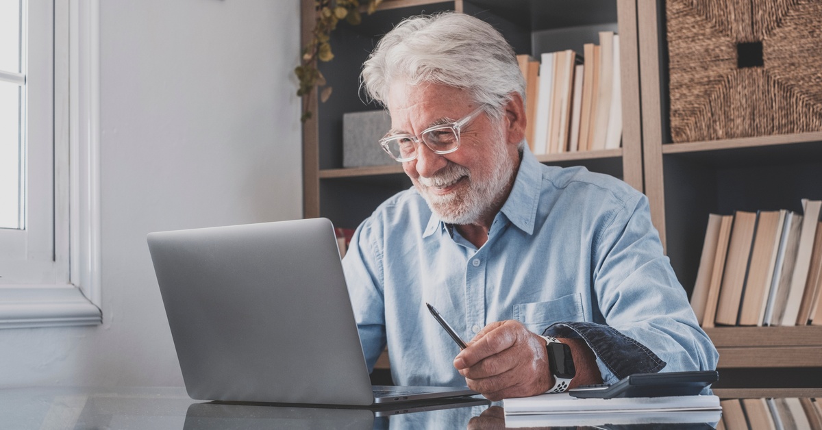 A person wearing glasses and holding a pen sits in front of a laptop and smiles while looking at the screen.